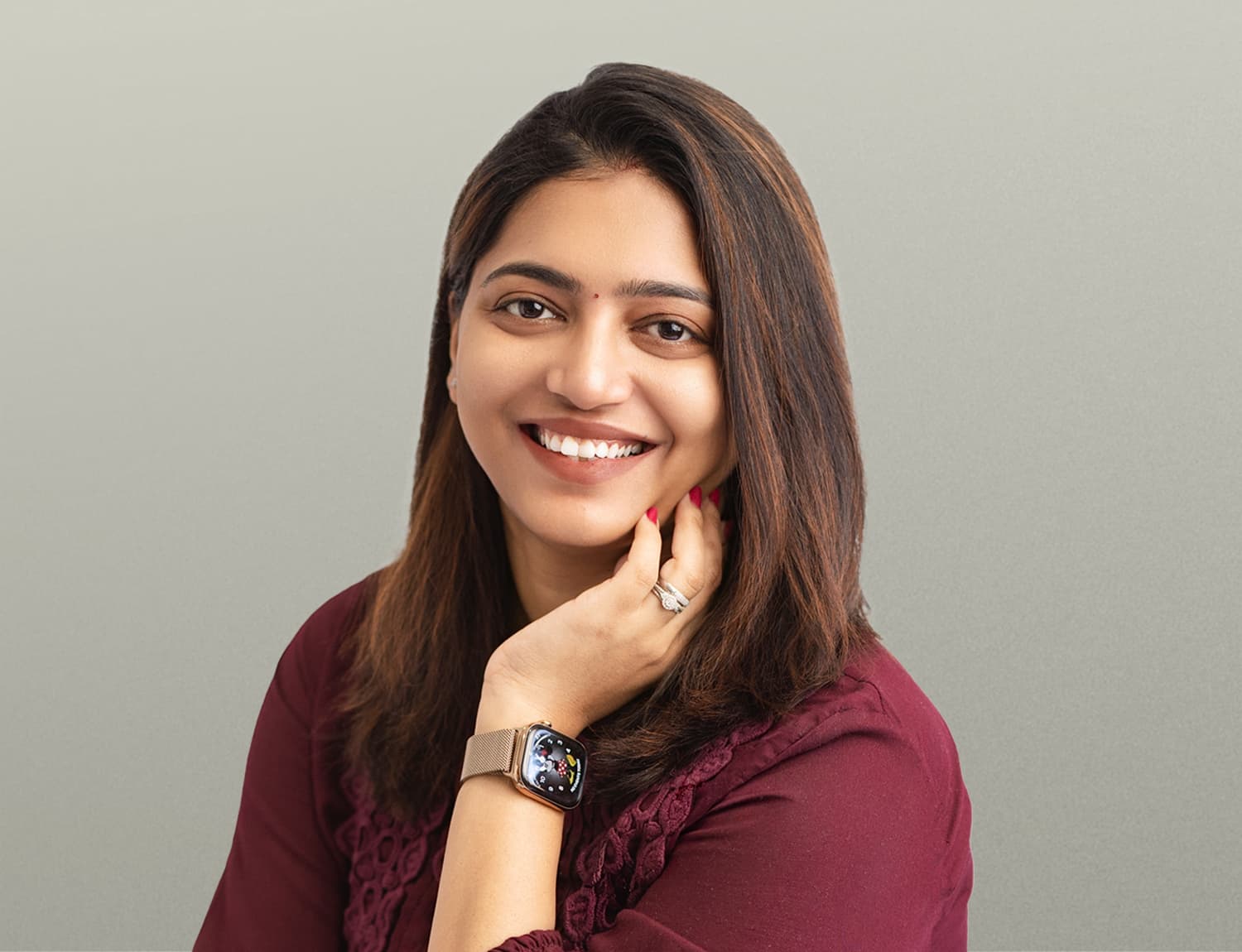 Professional portrait of Uma Kavuri, a woman with long brown hair wearing a burgundy blouse with lace detailing, smiling warmly while resting her hand thoughtfully under her chin, wearing a smartwatch with a metal band and red nail polish against a neutral beige background