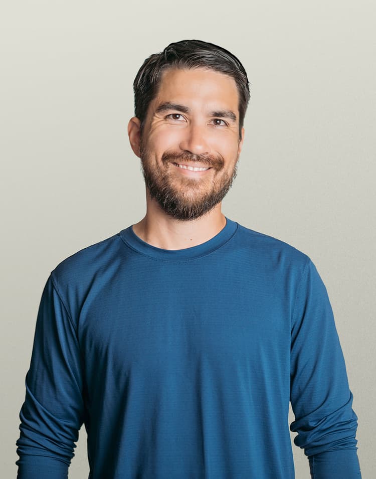 Professional headshot of Chris Devidal, a man with dark hair and a full beard wearing a blue long-sleeved shirt, smiling warmly at the camera against a neutral beige background
