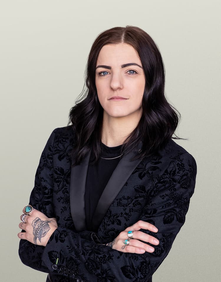 Professional headshot of Maranda Milia, a woman with dark wavy hair wearing a black textured blazer with arms crossed confidently, looking directly at the camera with a slight smile against a neutral beige background