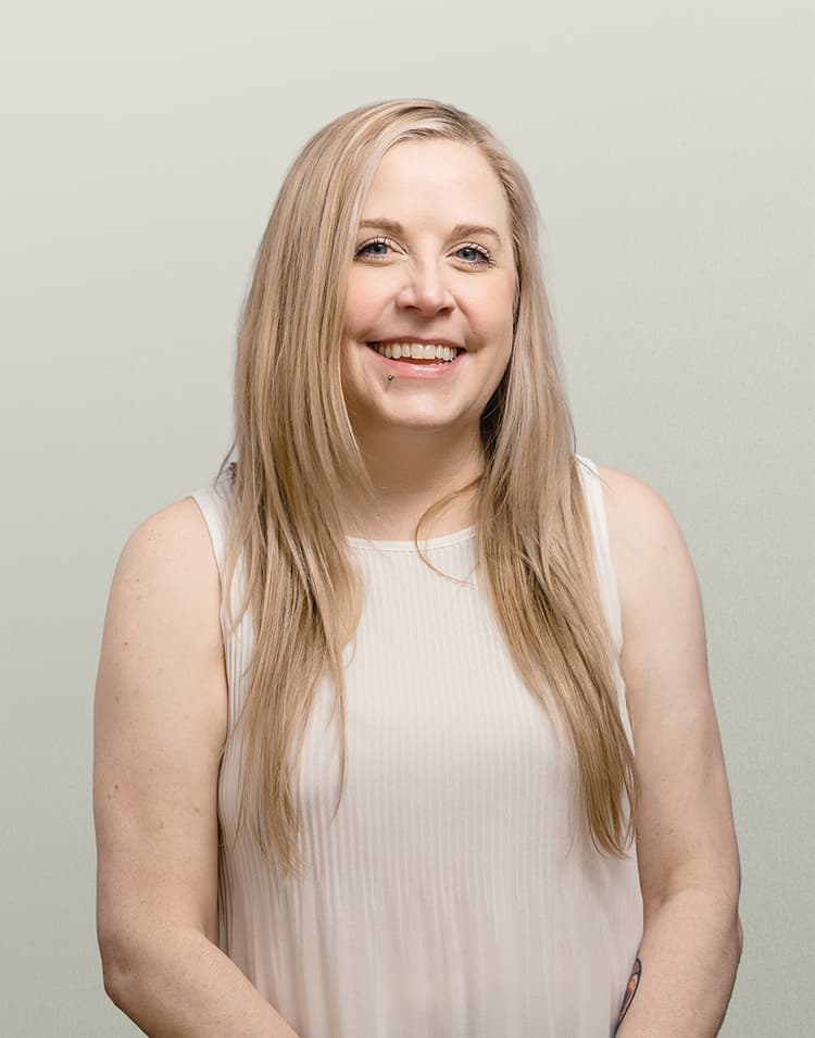 Professional headshot of Stacy Spakowsky, a woman with long blonde hair wearing a light pink sleeveless top, laughing joyfully at the camera against a neutral beige background