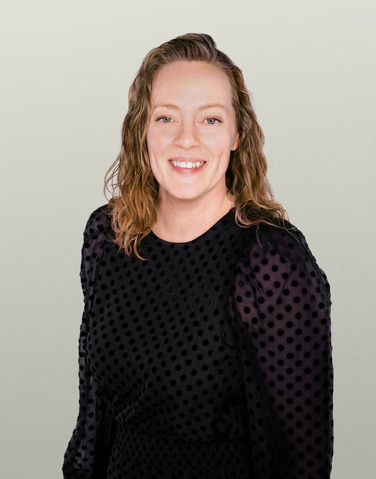 Professional headshot of Tami Holms, a woman with shoulder-length wavy brown hair wearing a black top with polka dot detailing, smiling brightly at the camera against a neutral beige background